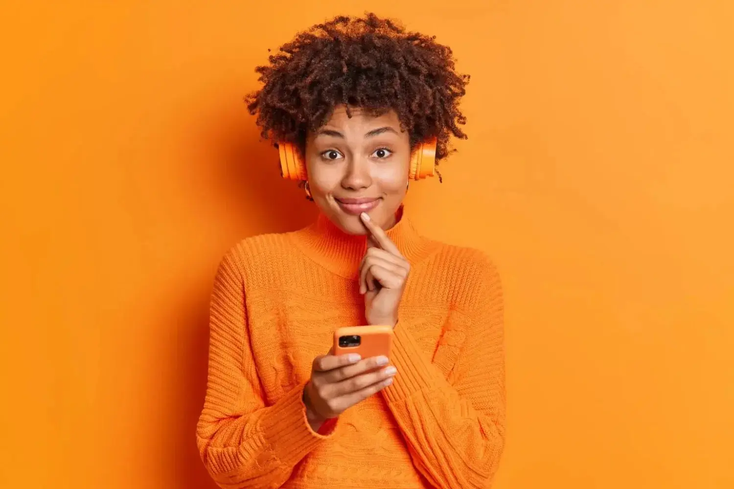 pretty-satisfied-woman-smiles-gently-front-keeps-finger-near-lips-uses-smartphone-online-texting-listening-music-wears-stereo-wireless-headphones
